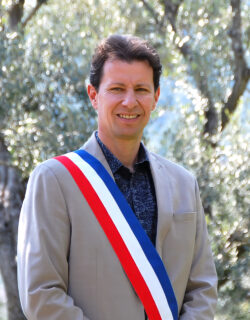 Photo d’Yves Palmieri, maire de La Farlède, portant une écharpe tricolore (bleu, blanc, rouge), debout devant un olivier en extérieur.