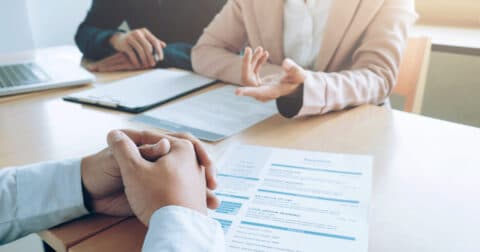 "Photo d'un entretien professionnel où une personne tend la main vers un CV posé sur une table. Une autre personne, assise en face, écoute attentivement. Un ordinateur portable et un bloc-notes sont également visibles sur la table."