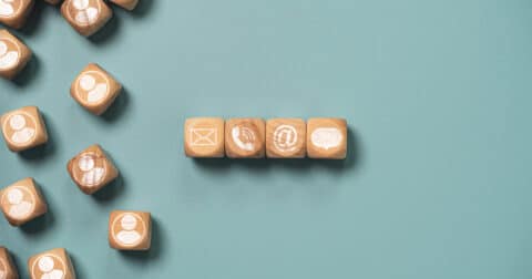 "Photo de cubes en bois disposés sur un fond bleu clair. À gauche, des cubes avec des icônes de profils humains. À droite, une ligne de cubes représentant des icônes de communication : email, appel téléphonique, notification et bulle de discussion."