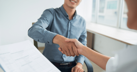 "Photo de deux personnes en train de se serrer la main à travers un bureau, symbolisant un accord, une entente ou une poignée de main professionnelle. L'une des personnes porte une chemise bleue et sourit, tandis que l'autre, partiellement visible, tend la main."