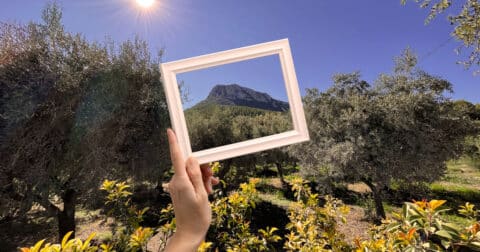 "Photo montrant une main tenant un cadre vide en plein air, encadrant une montagne verdoyante sous un ciel bleu et un soleil éclatant, avec des oliviers et des arbustes en premier plan."