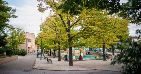 "Photographie d’un parc ou d’une aire de jeux publique en automne, avec des arbres aux feuilles jaunes et vertes. On y voit des bancs, une aire de jeux colorée pour enfants, des lampadaires, et un bâtiment en arrière-plan. Le sol est recouvert de sable et de feuilles tombées, sous un ciel partiellement nuageux."