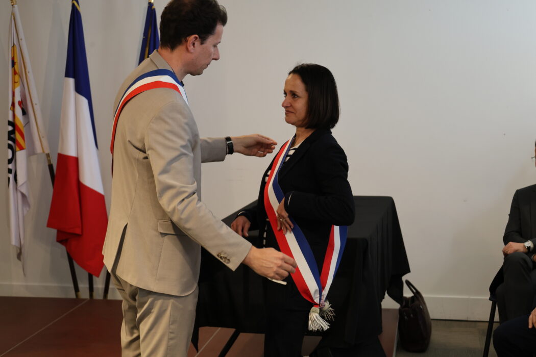 Photo de la remise d'écharpe du maire à sa première adjointe lors d'une cérémonie officielle. Le maire, portant une écharpe tricolore, ajuste l'écharpe similaire autour du cou de sa première adjointe, qui sourit avec émotion. Les drapeaux français et municipal sont visibles en arrière-plan.