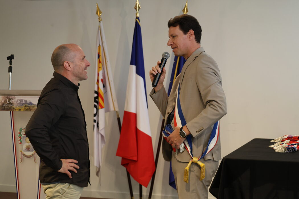 Photo de la remise d'écharpe à un adjoint par le maire lors d'une cérémonie officielle. Le maire, à droite, vêtu d'une écharpe tricolore, tend un micro à son adjoint, qui sourit et semble écouter attentivement. Les drapeaux français et municipal sont visibles en arrière-plan.