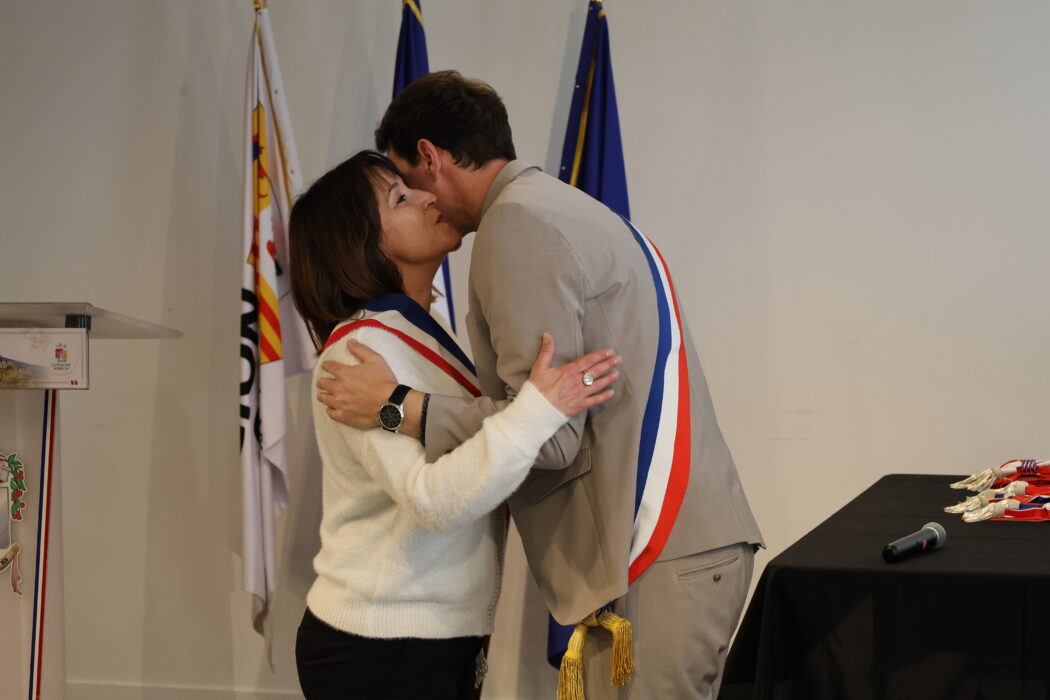 Photo d'une élue embrassant le maire lors d'une cérémonie officielle. Tous deux portent une écharpe tricolore. Les drapeaux français et municipal sont visibles en arrière-plan, ainsi qu'un pupitre décoré aux couleurs de la commune.