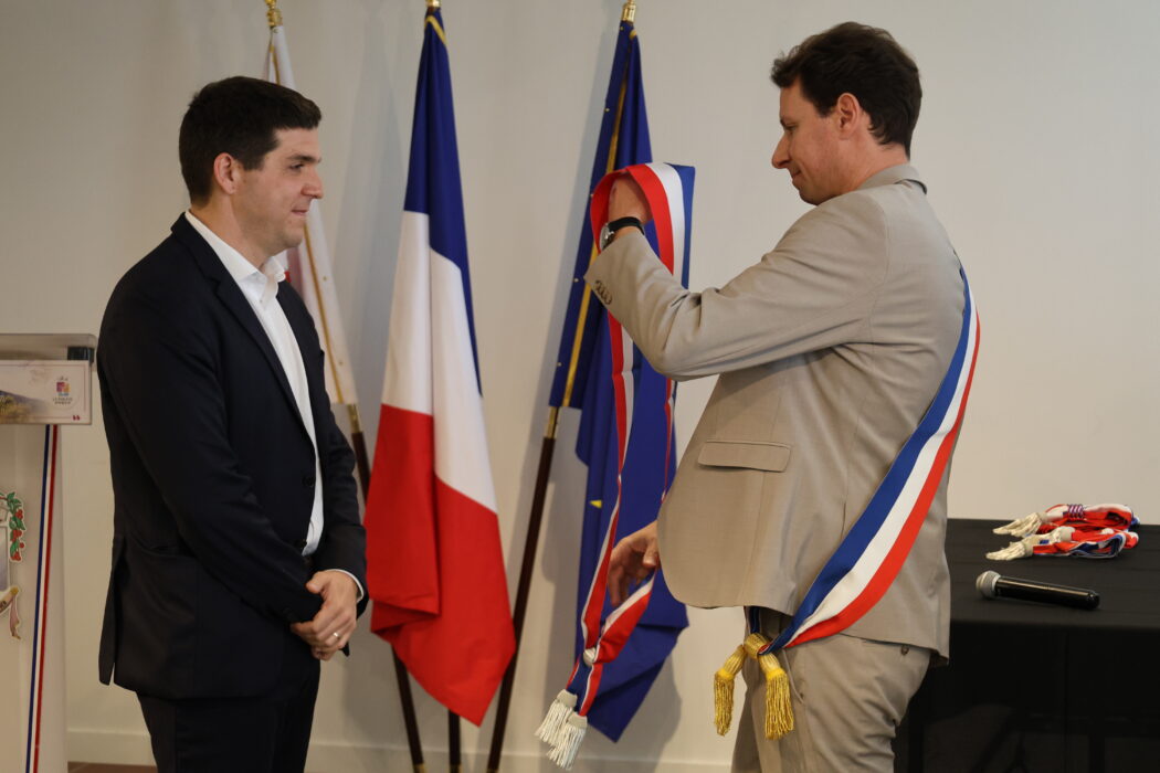 Photo du maire ajustant une écharpe tricolore autour du cou d'un adjoint lors d'une cérémonie officielle. Les deux hommes sont habillés de manière élégante, et les drapeaux français et européen sont visibles en arrière-plan.