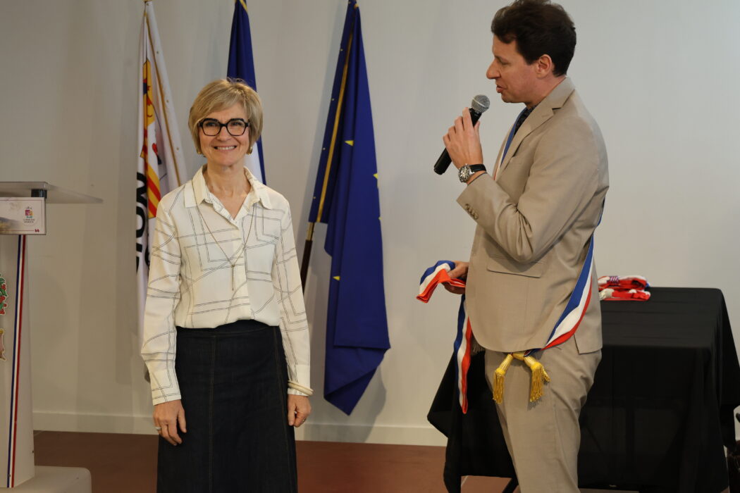 Photo du maire, vêtu d'une écharpe tricolore, en train de remettre symboliquement une écharpe à une élue souriante lors d'une cérémonie officielle. Les drapeaux français et européen sont visibles en arrière-plan, ainsi qu'un pupitre décoré aux couleurs de la commune.