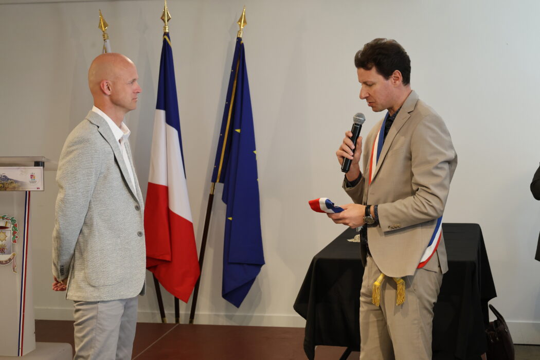 Photo du maire, vêtu d'une écharpe tricolore, en train de remettre une écharpe à un adjoint lors d'une cérémonie officielle. Le maire tient un micro et une écharpe tricolore, tandis que l'adjoint écoute attentivement. Les drapeaux français et européen sont visibles en arrière-plan.
