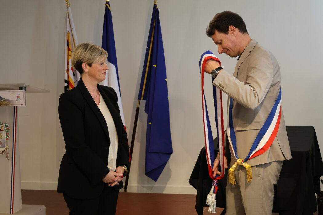Photo du maire, vêtu d'une écharpe tricolore, en train de remettre une écharpe à une élue lors d'une cérémonie officielle. L'élue, vêtue d'un tailleur noir et blanc, observe attentivement tandis que le maire ajuste l'écharpe. Les drapeaux français et européen sont visibles en arrière-plan.