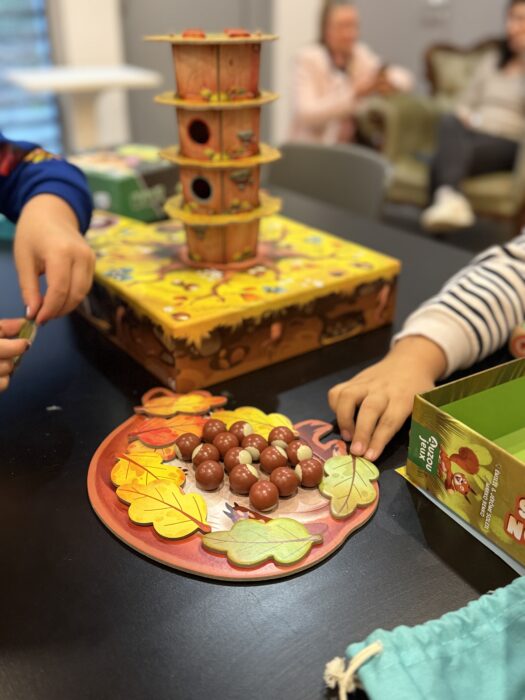 Photo d'un après-midi jeux de société à la médiathèque. Des enfants jouent à un jeu en bois coloré représentant un arbre avec des écureuils et des feuilles. Sur la table, des pièces en forme de glands et des feuilles en bois sont manipulées par les joueurs. L'ambiance est conviviale et ludique.