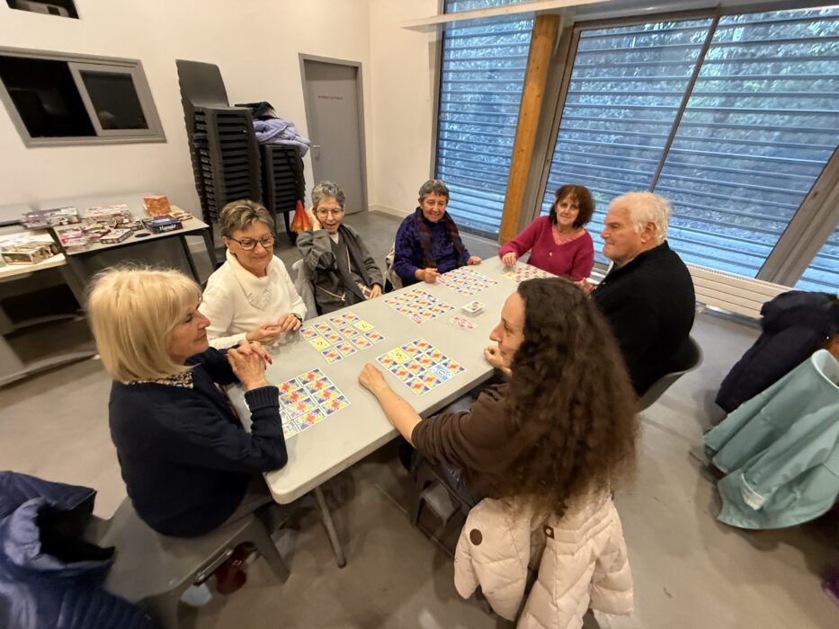 Photo d'un après-midi jeux de société à la médiathèque. Un groupe de personnes est assis autour d'une table, jouant à un jeu de cartes coloré. Les participants semblent concentrés et souriants, partageant un moment convivial. En arrière-plan, des étagères remplies de jeux de société sont visibles.
