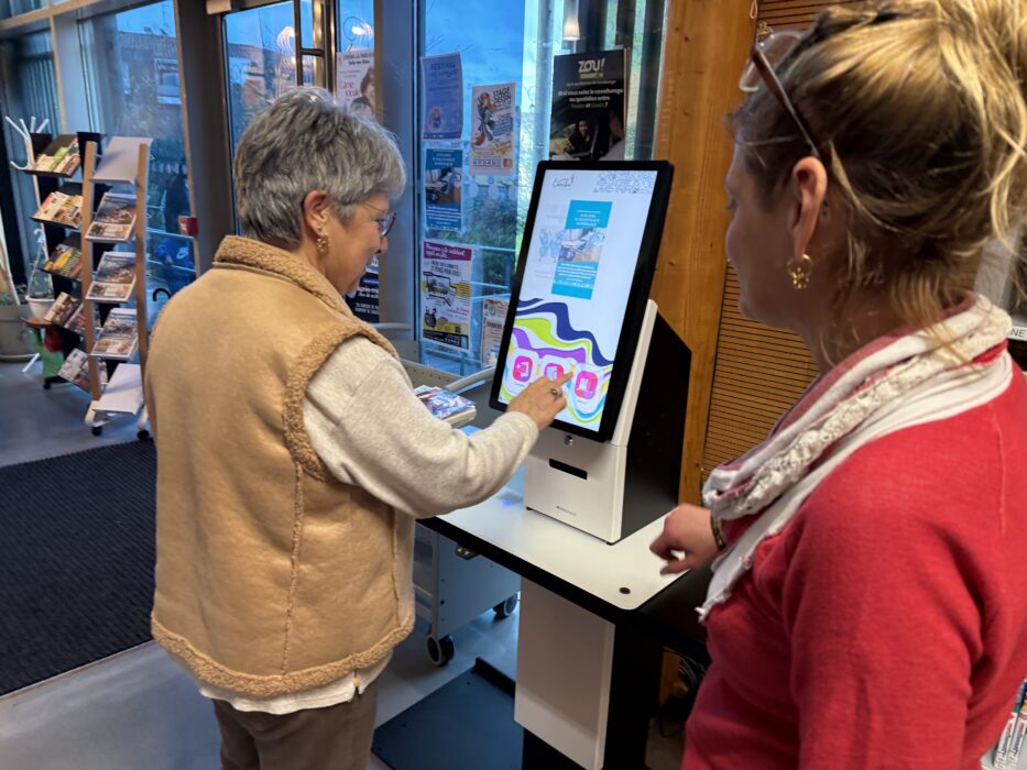 Photo de la nouvelle borne de retour de livres à la médiathèque. Deux personnes utilisent la borne tactile, qui affiche un écran coloré avec des instructions claires. L'espace est lumineux, avec des affiches et des présentoirs de brochures en arrière-plan.