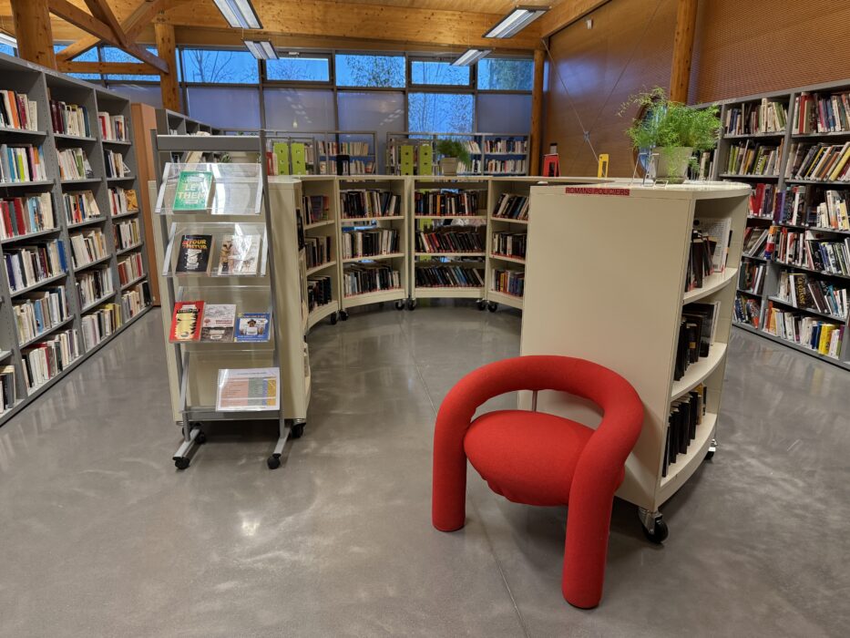Photo de l'espace réaménagé de la médiathèque Eurêka. Les étagères circulaires et linéaires sont remplies de livres variés. Un présentoir mobile avec des livres en évidence est visible au centre, entouré d'un fauteuil rouge design. Des plantes vertes et une lumière naturelle provenant des fenêtres en arrière-plan apportent une ambiance chaleureuse et accueillante