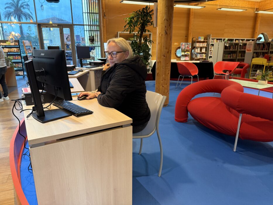 Photo du nouvel espace informatique de la médiathèque. Une personne utilise un ordinateur à un bureau moderne. Autour d'elle, des fauteuils rouges design et des plantes vertes créent une ambiance chaleureuse et conviviale. Les grandes fenêtres laissent entrer une lumière naturelle abondante.