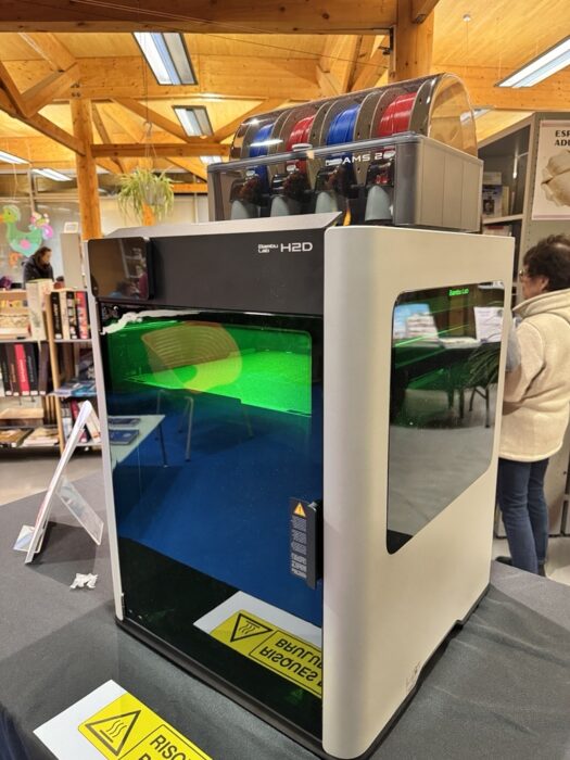 Photo d'une imprimante 3D Bambu Lab H2D installée dans la médiathèque. L'imprimante est en fonctionnement, avec un objet en cours d'impression visible à travers la vitre avant. Des bobines de filament de différentes couleurs sont placées sur le dessus. Des panneaux de sécurité indiquent les risques de brûlure et les précautions à prendre.