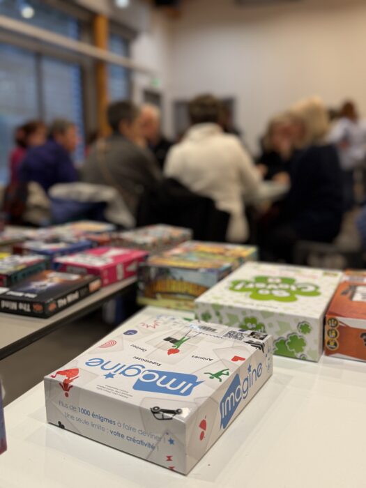 Photo d'un après-midi jeux de société à la médiathèque. Au premier plan, la boîte du jeu "Imagine" est visible, avec son design blanc et ses pictogrammes colorés. En arrière-plan, plusieurs autres jeux de société sont disposés sur une table, et des participants discutent et jouent, créant une ambiance conviviale et animée.