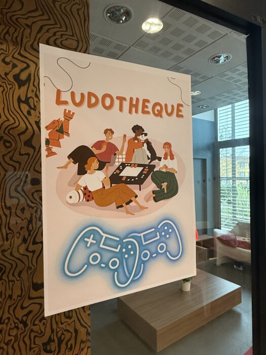 Affiche "Ludothèque" illustrée avec des personnages jouant à des jeux de société et une manette de jeu stylisée en bas. L'affiche est colorée et met en avant une ambiance conviviale et ludique.