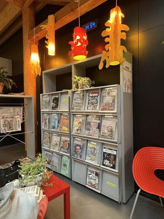 Photo de l'espace presse de la médiathèque. Un présentoir métallique expose divers magazines et revues, classés par catégories. Des luminaires design orange éclairent l'espace, et des plantes vertes apportent une touche naturelle. Une table rouge et des fauteuils colorés complètent l'aménagement.