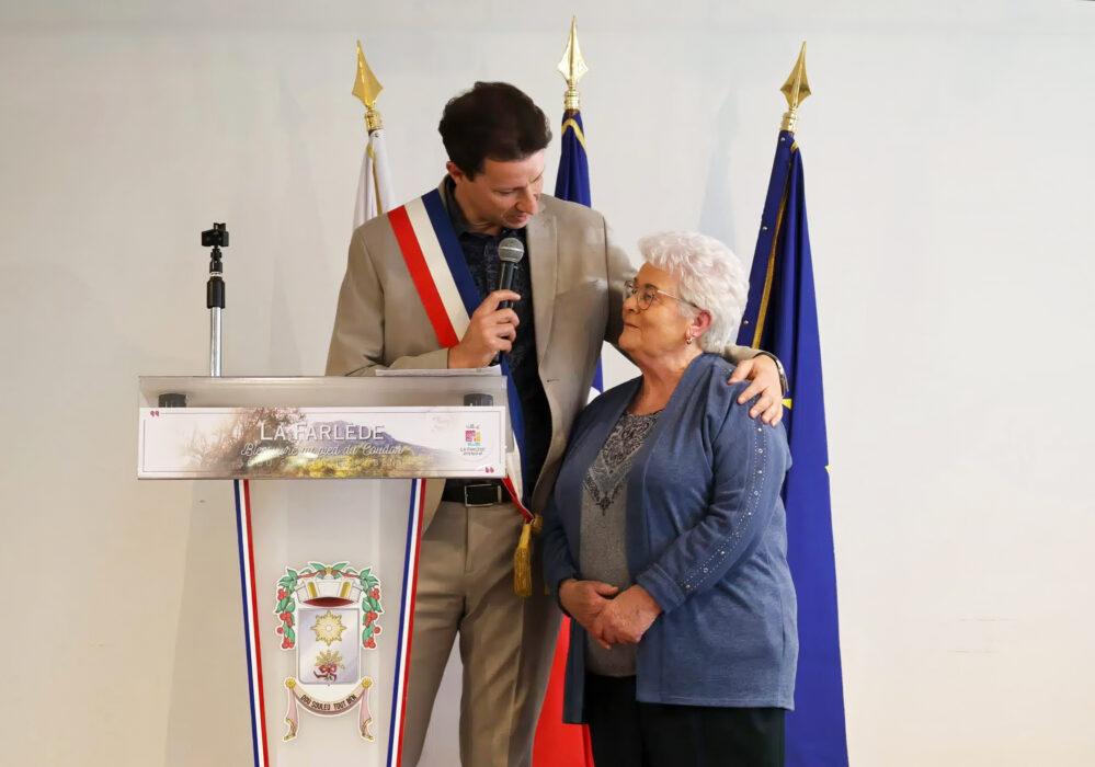 Photo d'Yves Palmieri, portant une écharpe tricolore, en train de s'adresser à Micheline Téobald lors d'une cérémonie officielle. Ils sont debout devant un pupitre orné du logo de La Farlède et des drapeaux français et européen.