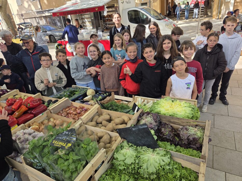 Les enfants du centre de loisirs posent fièrement devant un stand de légumes frais au marché lors du Printemps du Coudon. Derrière eux, des caisses remplies de poivrons, pommes de terre, salades, ail, oignons, et autres légumes de saison sont exposées. L’ambiance est ensoleillée et conviviale, avec des passants et des commerçants en arrière-plan.