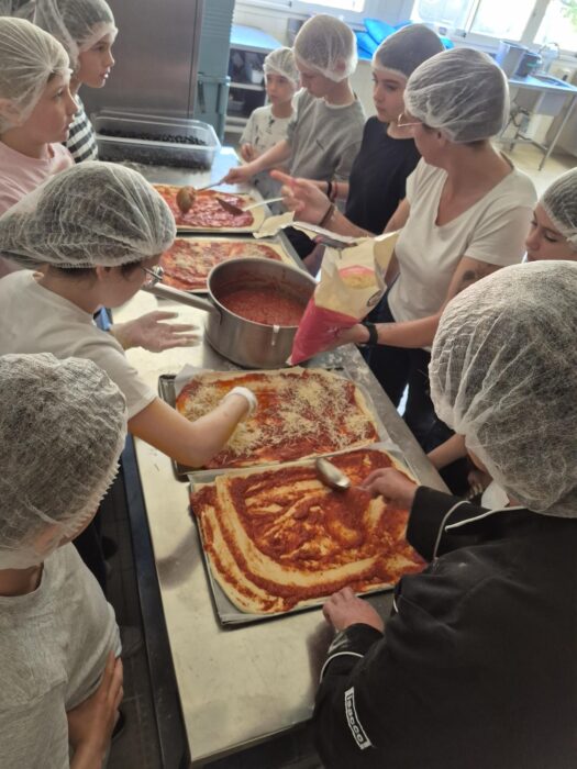 Atelier cuisine organisé par l’accueil de loisirs lors du Printemps du Coudon. Des enfants et des animateurs, vêtus de charlottes et de tabliers, préparent ensemble des pizzas. Ils étalent la sauce tomate et ajoutent les garnitures sur des pâtes à pizza posées sur des plaques. L’ambiance est conviviale et créative.