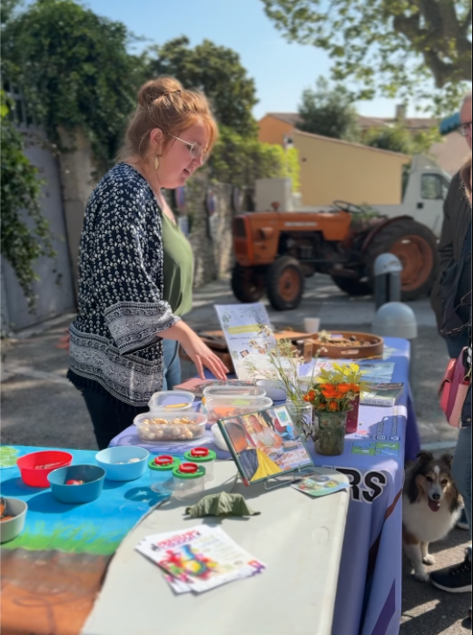 Stand animé lors du Printemps du Coudon. Une personne est penchée sur une table recouverte de documents, d’objets artisanaux, et de décorations florales. En arrière-plan, un tracteur et un chien ajoutent une touche rurale et chaleureuse à l’ambiance.