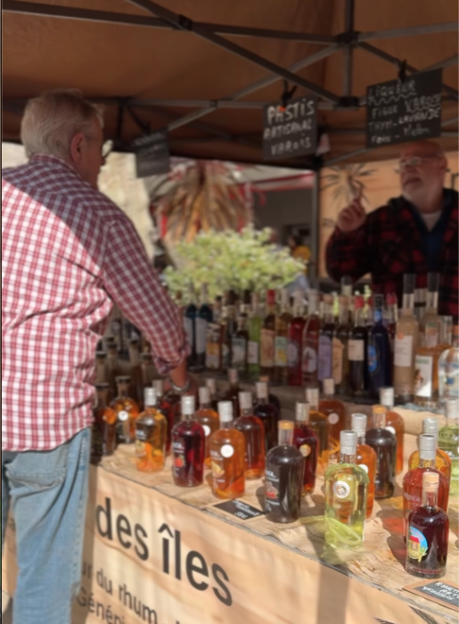 Stand de dégustation de rhums artisanaux lors du Printemps du Coudon. Un visiteur observe une table recouverte de nombreuses bouteilles de rhums variés, étiquetées et alignées. Un animateur se tient derrière le stand, entouré de plantes décoratives et d’ardoises indiquant les produits proposés.