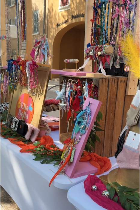 Stand de créations artisanales 'Ella Bella' lors du Printemps du Coudon. Une table recouverte d’une nappe blanche présente des colliers, des porte-clés colorés, des accessoires en tissu et des décorations suspendues. Les couleurs vives et les motifs variés attirent l’attention des visiteurs.