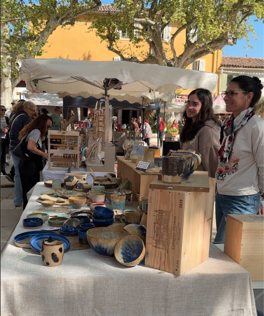 Stand de céramiques artisanales lors du Printemps du Coudon. Une table recouverte d’une nappe blanche expose des bols, des tasses et des plats en céramique aux motifs variés. Deux femmes discutent près du stand, entouré de caisses en bois et d’autres créations. En arrière-plan, des visiteurs explorent les autres stands sous des tentes.
