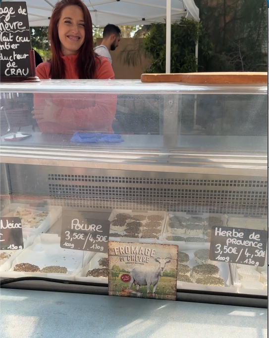 Stand de fromages lors du Printemps du Coudon. Une vendeuse souriante se tient derrière une vitrine réfrigérée présentant divers fromages de chèvre, dont du 'Poivre' et des 'Herbes de Provence'. Une ardoise indique les prix et un visuel de chèvre décore la vitrine.
