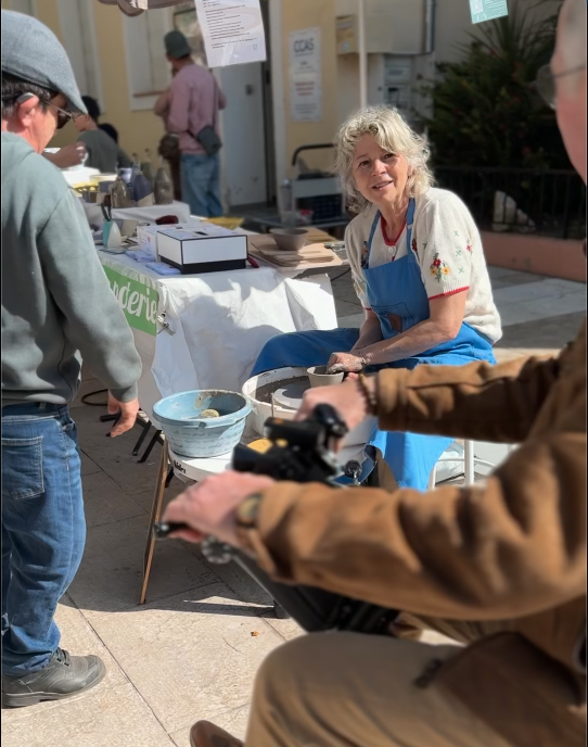 Artisane souriante assise à un stand de poterie lors du Printemps du Coudon. Elle porte un tablier bleu et un pull à motifs floraux, et tient un outil de poterie. Autour d’elle, des visiteurs observent et manipulent des créations en argile. En arrière-plan, une table avec du matériel et des affiches est visible.