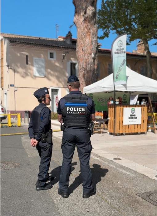 Deux policiers municipaux en tenue officielle, de dos, veillant à la sécurité lors du Printemps du Coudon. En arrière-plan, un stand de boissons 'Hop Green' avec une tente blanche et une signalétique visible. L’ambiance est ensoleillée, dans un cadre urbain avec des arbres et des bâtiments.
