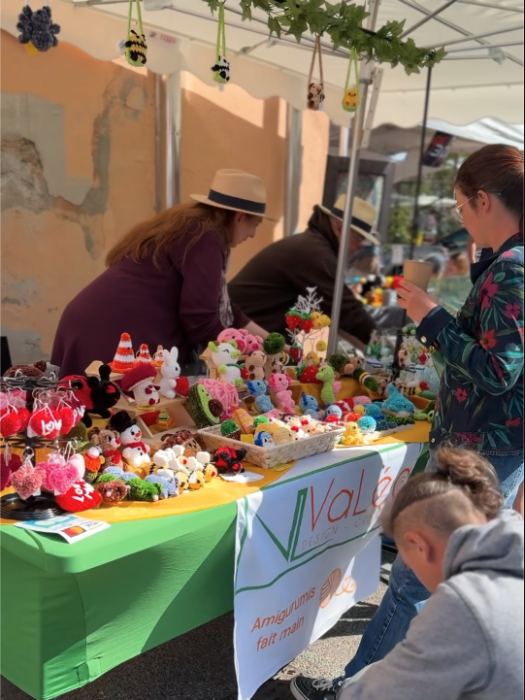 Stand d’amigurumis fait main lors du Printemps du Coudon. Plusieurs peluches et figurines colorées sont exposées sur une table recouverte d’une nappe verte et jaune. Deux femmes discutent près du stand, tandis qu’un enfant observe les créations. Une banderole indique 'Amigurumis fait main'.