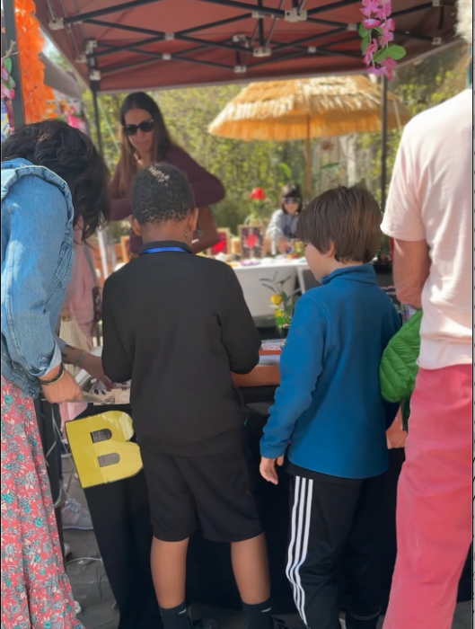 Groupe de personnes, dont des enfants, participant à une activité sous une tente décorée de guirlandes lors du Printemps du Coudon. Un enfant tient une lettre en carton jaune avec la lettre 'B'. En arrière-plan, une animatrice encadre l’activité, entourée de tables et de matériel créatif.
