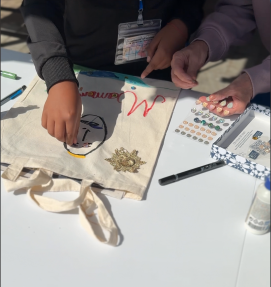 Atelier de personnalisation de tote bags lors du Printemps du Coudon. Une personne utilise un fer à dorures pour appliquer un motif sur un sac en tissu blanc, tandis qu’une autre main tient le sac. Sur la table, des autocollants dorés et des accessoires de décoration sont visibles.