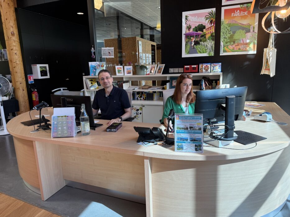 Banque d’accueil de la médiathèque de La Farlède, avec deux agents souriants assis derrière un comptoir en bois clair. Des écrans, des documents informatifs, et des affiches colorées représentant la commune sont visibles. L’espace est lumineux et organisé, avec des étagères remplies de livres et de ressources en arrière-plan.