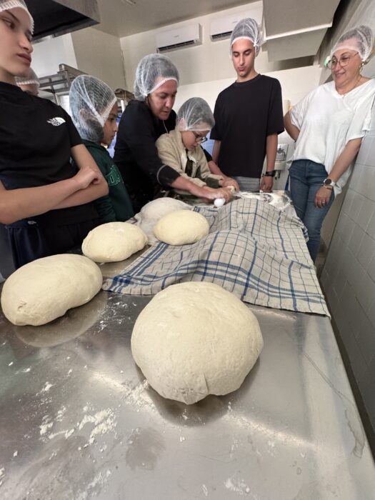 Atelier de boulangerie au centre de loisirs : des enfants et des adultes, vêtus de charlottes, façonnent des pâtons de pain sur une table farinée. Plusieurs boules de pâte sont alignées sur un torchon à carreaux, sous la supervision attentive des animateurs. L’ambiance est collaborative et éducative."