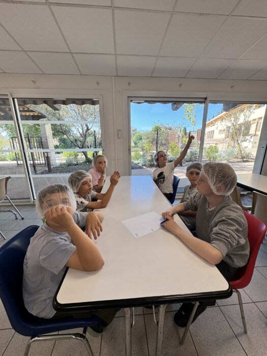 Enfants du centre de loisirs assis en salle avec des charlottes, participant à un atelier culinaire. Ils sont attablés et écoutent attentivement une animatrice. L’un d’eux lève la main pour poser une question, tandis que les autres semblent concentrés. En arrière-plan, une baie vitrée laisse entrevoir un espace extérieur ensoleillé.