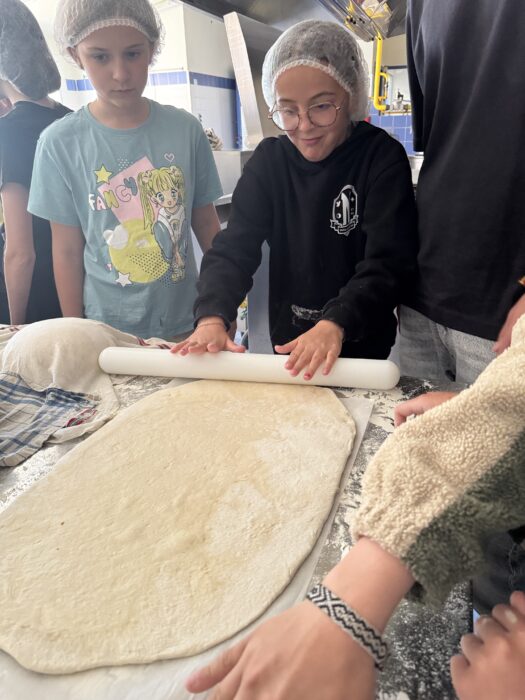 Atelier de cuisine au centre de loisirs : des enfants vêtus de charlottes étalent une pâte à pizza sur un plan de travail fariné, sous la supervision d’un adulte. L’un des enfants utilise un rouleau à pâtisserie pour aplatir la pâte, tandis que les autres observent et participent activement.