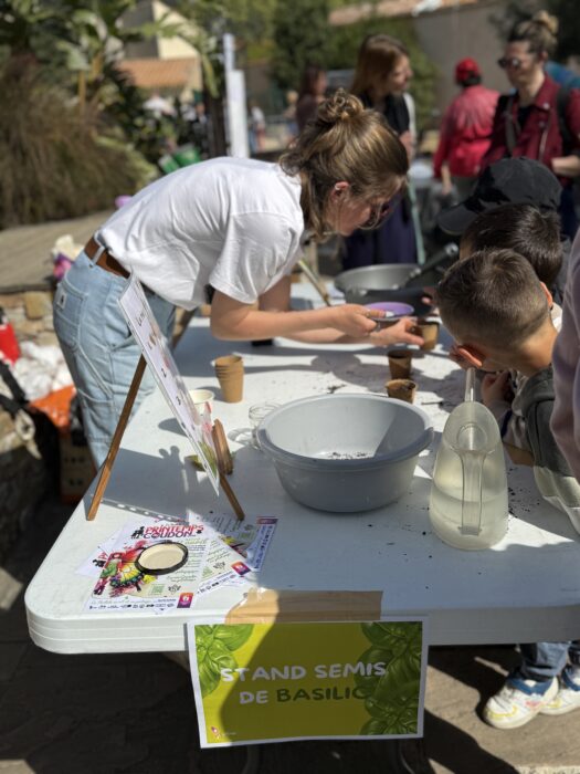 Atelier de semis de basilic lors du Printemps du Coudon. Une animatrice aide des enfants à remplir des petits pots de terreau et à y planter des graines de basilic. Sur la table, un bol de terreau, une carafe d’eau, des pots en plastique, et une affiche 'Stand Semis de Basilic' sont visibles. L’ambiance est conviviale et éducative.