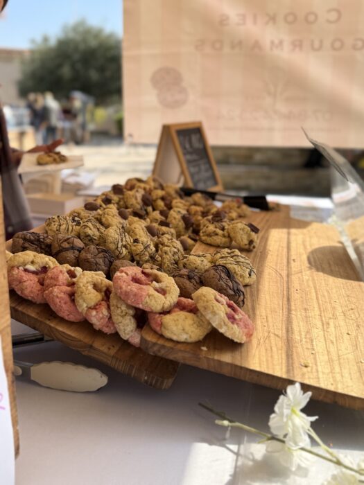 Plateau en bois garni de cookies variés, exposés lors du Printemps du Coudon. Les cookies, de différentes couleurs et saveurs, sont soigneusement disposés et prêts à être dégustés. En arrière-plan, une enseigne indique 'Cookies Gourmands', et une fleur blanche ajoute une touche décorative.