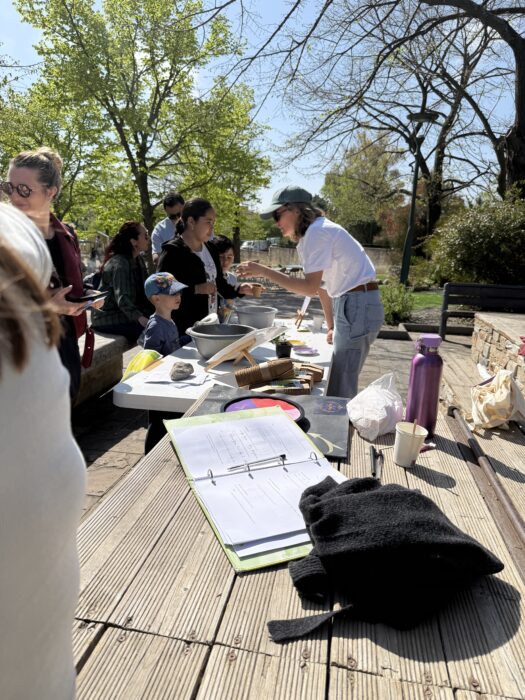 Atelier participatif en extérieur lors du Printemps du Coudon, où des enfants et des adultes sont réunis autour d’une table recouverte de bols, d’outils, et de matériaux naturels. Une animatrice guide les participant·e·s dans une activité créative ou culinaire. L’ambiance est conviviale, sous un ciel ensoleillé et entouré de verdure