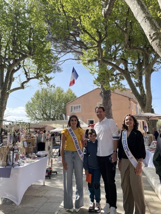Le maire de La Farlède entouré des Miss La Farlède 2026, posant devant des stands artisanaux lors du Printemps du Coudon. Les Miss portent leurs écharpes officielles, et un drapeau français flotte en arrière-plan. L’ambiance est ensoleillée, avec des arbres et des visiteurs autour des stands