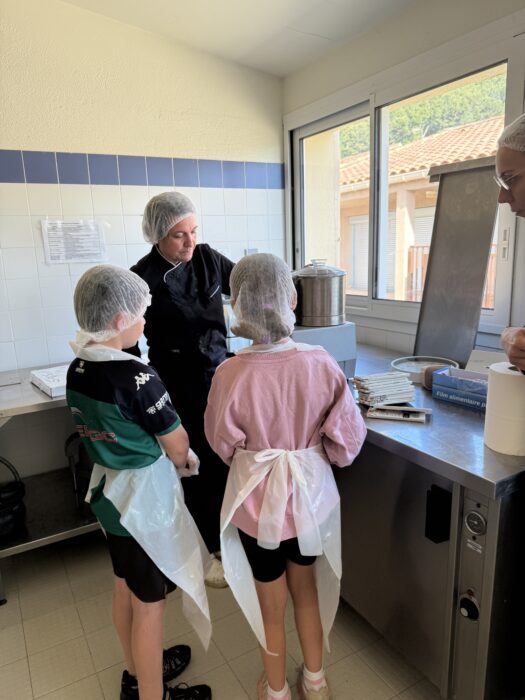 Atelier culinaire au centre de loisirs : une animatrice en tenue professionnelle encadre deux enfants vêtus de charlottes et de tabliers, leur montrant comment utiliser un équipement de cuisine. Les enfants écoutent attentivement les consignes, dans une cuisine lumineuse et bien équipée.