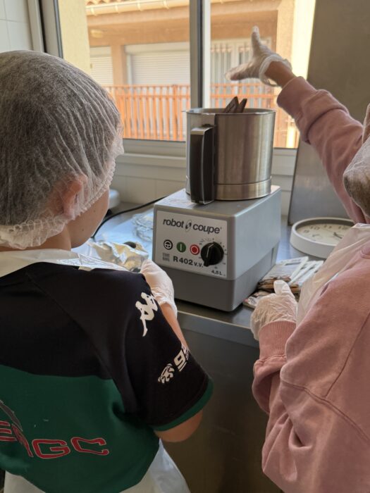 Atelier culinaire au centre de loisirs : deux enfants, vêtus de charlottes et de gants, utilisent un robot coupe pour préparer une recette. L’un d’eux montre du doigt le bol du robot, tandis que l’autre observe attentivement. L’appareil est posé sur un plan de travail près d’une fenêtre ensoleillée.