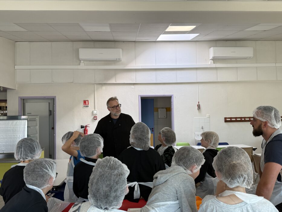 Atelier culinaire au centre de loisirs : un groupe d’enfants et d’adultes, vêtus de charlottes et de tabliers, écoutent attentivement les consignes données par un animateur. Ils sont assis autour de tables dans une cuisine collective équipée. L’ambiance est studieuse et collaborative.