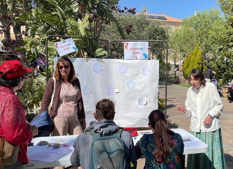 Animation organisée par les ambassadeurs de la transition sur la manière de repenser la ville lors du Printemps du Coudon. Une animatrice discute avec des enfants et des adultes autour d’un panneau où sont apposés des autocollants et des post-it. En arrière-plan, des visiteurs déambulent dans un parc verdoyant et ensoleillé.