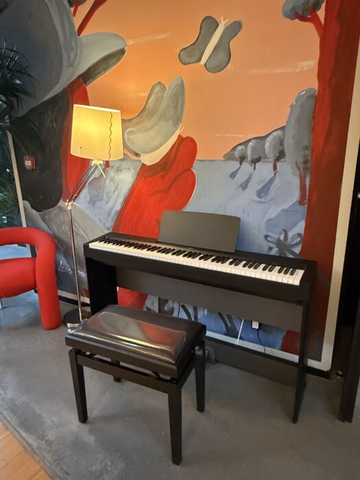 Piano électrique noir installé dans un espace coloré de la médiathèque de La Farlède. Il est accompagné d’un tabouret réglable et se trouve à proximité d’une lampe design et d’un fauteuil rouge. En arrière-plan, une fresque murale artistique apporte une touche créative à l’espace.
