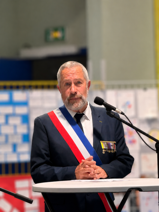 Cérémonie de transmission du drapeau Unadif au collège André-Malraux de La Farlède. Le conseiller municipal correspondant Défense, vêtu d’un costume sombre et portant l’écharpe tricolore, s’exprime au micro. Il arbore plusieurs médailles sur sa veste, symbolisant son engagement. Ses mains sont jointes devant lui, et il semble s’adresser à l’assistance avec gravité."
