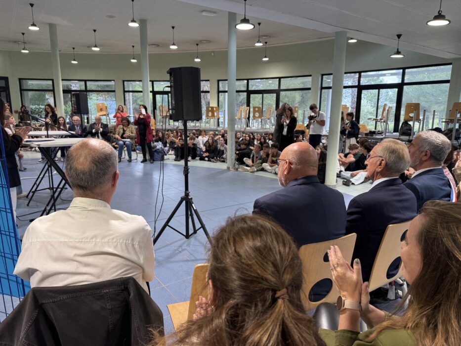 Cérémonie de transmission du drapeau Unadif au collège André-Malraux de La Farlède. Un public composé d’élèves et d’adultes est assis dans une salle lumineuse avec de grandes baies vitrées. Sur scène, des intervenants s’expriment au micro, tandis que certains participants applaudissent. L’ambiance est solennelle et collective, avec une attention particulière portée aux discours.